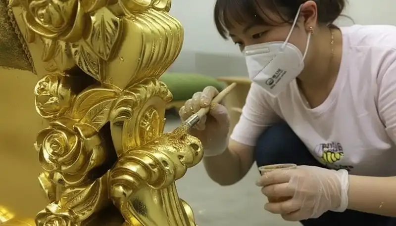 A young East Asian woman with light skin is diligently applying gold leaf or gold paint to an ornate, baroque-style golden object. She is positioned in the lower-right quadrant, kneeling or crouching, with her body angled slightly toward the object. She appears to be in her 20s or 30s. Her dark hair is pulled up, and she is wearing a white t-shirt, dark trousers, and disposable clear plastic gloves on both hands. She is also wearing a white protective N95 or similar face mask. In her right hand, she holds a small, light-colored wooden brush, which she is using to apply the gold material to a detailed, curved portion of the object, which features elements resembling carved roses and scrolls. In her left hand, she holds a small, clear plastic container containing the gold material, which appears granular or flaky, possibly gold powder or gold leaf flakes suspended in a medium. The golden object dominates the left and central parts of the image and is highly detailed with sculpted floral and scrolling motifs. The background is slightly blurred and includes soft, muted colors, suggesting an indoor workshop or studio environment, with a hint of a green or dark cushion visible behind the woman. The lighting is bright, illuminating the process and highlighting the reflective quality of the gold finish.